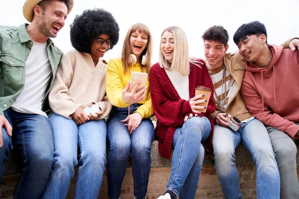 Group of multicultural young teenagers sitting together looking phone and laughing out loud. Gathering of friends to have fun watching entertainment content on the cell. Addicted to social networks.