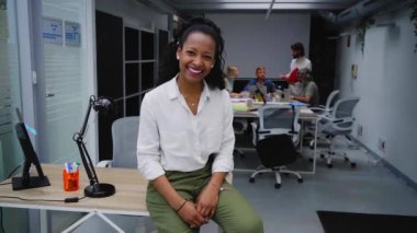 Video of smiling young African American professional female sitting at modern office desk. Happy mixed race business woman team leader posing for photo at corporate meeting. Concept of workplace. 