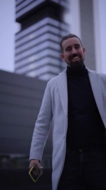 Vertical video handsome young successful businessman celebrates dancing a new professional success outside a business center. Close-up of ceo in overcoat smiling happily at camera outdoors. 