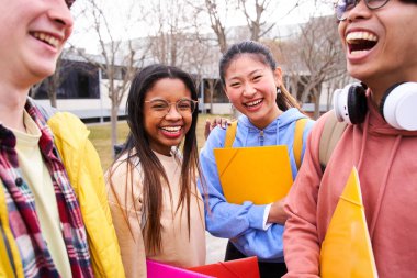 Portrait of four international students with beaming smiles posing for photo. Focus on the two cheerful young girls holding folders. Multiracial people gathered outdoor in the school library. Autumn.