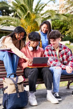 Vertical young international college students reading books, studying on laptop, preparing for exam or working on group project while sitting on bench of college campus university. People outdoors.