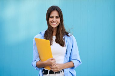 Portrait on blue background of smiling and cheerful young Caucasian girl. Pretty student woman holding yellow folder. Happy college people posing and looking at camera. Copy space.