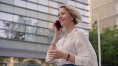 Close-up of a smiling blonde middle-aged Caucasian business woman talking on cell phone while walking to her workplace. Lawyer taking a call on the street in the city. Professional people outdoors.