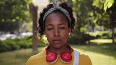 Video of beautiful African woman concentrating looking camera. Close up people in park outdoor. Student girl with headphones with serious look on face. Confident and thoughtful young college female.