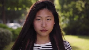 Close up of serious young Asian woman looking camera. Cute Chinese standing in city lifestyle street. Beautiful girl worried outdoors. Concentrated lady emotional expression on urban background. 