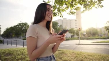 Smiling woman happily chatting on cell phone with friends and laughing. Beautiful and cheerful people outdoors. Attractive young girl using dating app while waiting in city park. Female empowered. 