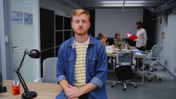 现代办公室里年轻的红头发程序员或商人严肃地看着摄像机 手交叉的自由人摆好姿势 集中在会议室拍照 同事们在幕后工作 — 图库视频影像