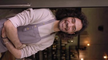 Vertical video young adult local small business owner looking at camera. Happy male bartender with positive attitude. Cheerful worker smiles while standing leaning on bar door. Confident people. 