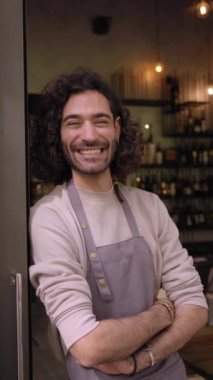 Vertical video young adult local small business owner looking at camera. Happy male bartender with positive attitude. Cheerful worker smiles while standing leaning on bar door. Confident people. 