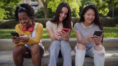 Three happy women using cell phone outdoors. Group smiling college friends checking social networks on smartphone. Classmates having fun watching something on the mobile. Young girls flirting online. 
