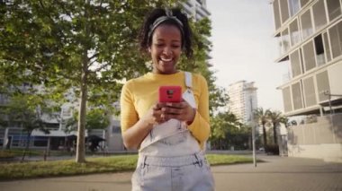 Gülümseyen Afro-Amerikan kadın cep telefonuyla arkadaşlarıyla sohbet ederken gülen beyaz dişler. Dışarıda neşeli insanlar var. Genç kız şehir parkında yürürken flört uygulaması kullanıyor..