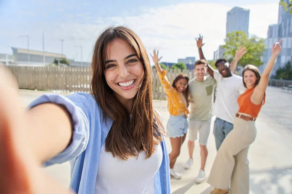 POV selfie outside of a smiling and cheerful pretty young girl with her friends in the background. Happy people looking camera and celebrating with hands up. Multiracial portrait college classmates.
