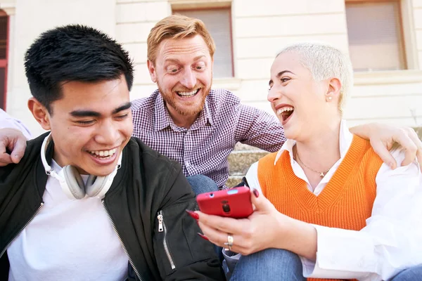Big laugh of a woman with multiracial friends using mobile. Three happy colleagues smiling funny sitting outdoor holding and looking at smart phone. People entertained with social networks.
