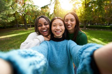 Dışarıdaki fotoğraf için poz veren üç gülümseyen kız, kameraya bakıyor. Üniversiteli kadınlar parkta selfie çekip sarılıyorlar. Çoklu etnik gruplar arasında dostluk ve eğlence ilkbahar toplantısı. 