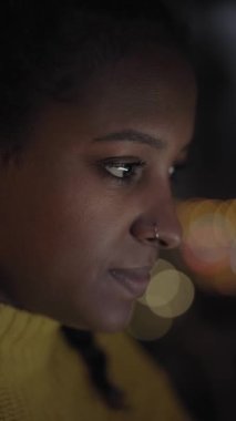 Evdeki karanlık odada, çok ırklı, şirin, siyah bir kızın dikey yakın portresi. Dizüstü bilgisayarına bakan konsantre bir kadın. İnsanlar arka planda odak dışı şehir ışıkları olan kapalı yerlerde çalışıyorlar..