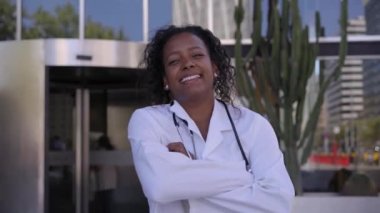 Steteskoplu laboratuvar önlüklü Afro-Amerikalı kadın doktor. Gülümseyen genç insanların tıbbi uzmanlık alanı açık havada poz vermeleri. Kolları bağlı mutlu ve neşeli bir insan..