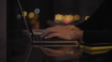 Evinin karanlık odasında laptopunu kullanan tanınmayan genç bir adamın ellerini kapatın. Bilgisayarda daktilo eden iş adamları. Şehir ışıkları arka planda bulanık bir şekilde kapalı alanda çalışan kişi. 