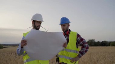 Sert şapkalı iki mühendis, tarımsal alandaki rüzgar türbinli planlara bakıyor. Profesyonel teknisyenler rüzgar santrali dışında konuşuyorlar. Temiz yenilenebilir enerji, sürdürülebilir gelecek.
