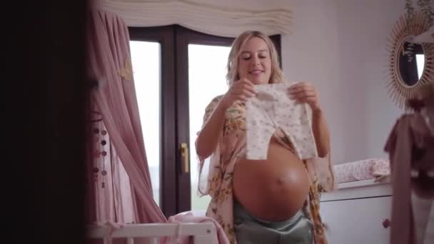 怀孕的高加索妇女为规划新生儿准备折叠的婴儿衣服 在孩子们的房间里 妈妈在等待着孩子的出生 一个新家庭成员的未来 产妇概念 — 图库视频影像
