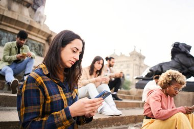 Group of individualistic people using phone outdoors with serious face. Friends focused on their mobiles sitting on a staircase in a city. Technology addicts concept. High quality photo