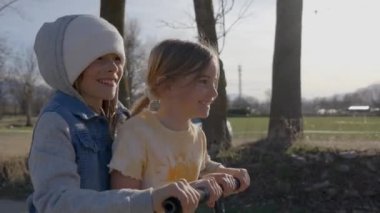Gülümseyen sarışın kardeşler açık hava parkında birlikte scooter kullanırlar. Çocuklar güneşli bir günde cadde boyunca tekerlek sürerken eğleniyorlar. Mutlu çocuklar aile aktivitelerinden zevk alırlar..