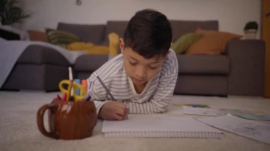 Latin küçük çocuk ödevlerine konsantre oluyor ve evin zeminine not defterine yazıyor. Çocuklar boş vakitlerinde oturma odasındaki halıya uzanmış çizim yapıyorlar. Evde eğitim ve çocuk gelişimi kavramı.