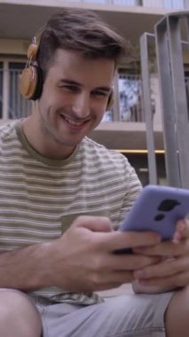 Hücre kullanan ve üniversite kampüsünün merdivenlerinde oturup gülen mutlu bir erkek öğrencinin dikey videosu. Gülümseyen, müzik dinleyen, mesajlaşan ve cep telefonundan sosyal ağları kontrol eden genç bir çocuk.. 