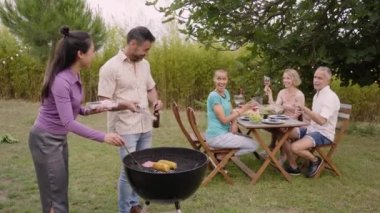 Olgun bir çift arka plandaki masada oturan ve tezahürat yapan arkadaşları için barbekü yapıyor. Meslektaşlar evdeki bahçede başarıyı kutluyorlar. Açık hava gündüz partisi. 