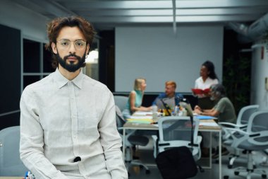 Meslektaşları arka planda farklı projeler üzerinde çalışırken kendinden emin genç erkek liderin oturduğu ve kameraya baktığı portre. Ciddi bir iş adamı. Çalışkan ve girişimci bir adam.. 