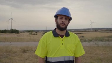 Büyük rüzgar türbinlerinin çalışmasını denetlemek için Doğal Enerji Rüzgâr türbini tesisinde çalışan genç mühendis elektrik gücünü kontrol ediyor. Erkek teknisyen kameraya ciddi ve kendinden emin bir şekilde bakıyor..