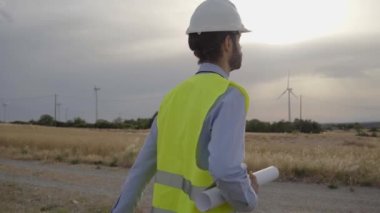 Teknik mühendis rüzgar çiftliğinin planlarını elinde tutarak yürüyor. Sürdürülebilir yenilenebilir enerji konusunda yetenekli bir işçi. Elektrik üretim istasyonu. Profesyonel teknisyen açık hava iş üniforması..