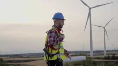 Teknik mühendis rüzgar tarlasında elinde telsizle dolaşıyor. Sürdürülebilir yenilenebilir enerji konusunda yetenekli bir işçi. Elektrik üretim istasyonu. Profesyonel teknisyen açık hava iş üniforması..