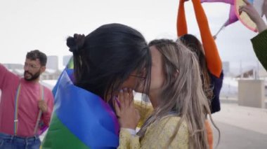 Eşcinsel gururu festivalinde öpüşen iki genç kız. Igbt partisini arka planda gökkuşağı bayrağı ve hayranlarıyla kutlayan bir grup arkadaş. Z nesli ve cinsellik türleri.