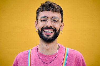 Gökkuşağı lgbt pantolon askılarını tutan neşeli, genç beyaz bir adam. Fotoğraf sarısı arka plan için poz veren eşcinseller. Neşeli eşcinsel sakallı insan izole edilmiş mutlu stüdyo fotoğrafı. Millennials.