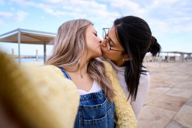 Mutlu LGBT genç lezbiyen beyaz çift dışarıda cep telefonuyla selfie çekmek için turist tatilinde öpüşüyor. Aşık eşcinseller ve romantik davranışlar. Sosyal ağlarda z nesli ve fotoğraflar. 