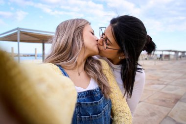 Happy LGBT young lesbian Caucasian couple on tourist vacation kissing for selfie with cell phone outdoors. Gay people in love and romantic attitude. Generation z and photos on social networks.  clipart