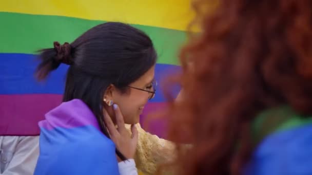 ゲイのプライドフェスティバルの日に屋外でキスをする2人の若いレズビアン女性 虹の旗やファンを背景にLgbtパーティーを祝う友人のグループ 世代Zとセクシュアリティの種類 — ストック動画