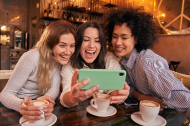 Üç genç arkadaş kafeteryada içki içerken cep telefonundan komik videolar izliyorlar. Gülümseyen mutlu kızlar birlikte zaman geçirip kahve içiyorlar. Milenyum Kadınları Sosyal Ağ.