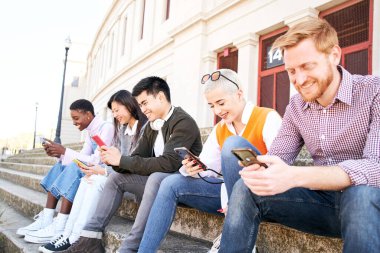 Çok ırklı bir grup arkadaş cep telefonlarını açık havada birlikte kullanıyor ve cep telefonlarını izlerken eğleniyorlar. Sosyal ağlara bağımlı gülümseyen gençlerin toplanması. Çevrimiçi iletişim yeni nesiller 