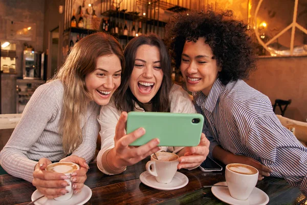 Üç genç arkadaş kafeteryada içki içerken cep telefonundan komik videolar izliyorlar. Gülümseyen mutlu kızlar birlikte zaman geçirip kahve içiyorlar. Milenyum Kadınları Sosyal Ağ.