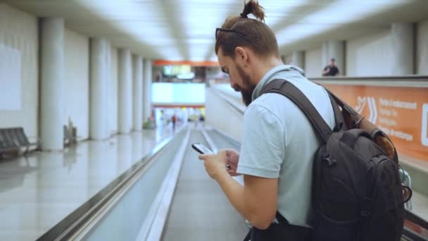 クールで現代的な空港ターミナルで若いヒップスター男性を探しています オンライン搭乗券やキャンセル出発 到着便のためのスマートフォンを使用してください 都市ノマド旅行ブロガー — ストック動画