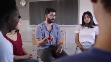 Çeşitli üniversite psikoloji öğrencileri bir grup sandalyede oturup bir çember oluşturuyorlar ve bir ders sırasında etkileşime giriyorlar. Öğretmen bir dizi soru için elmayı uzatır.. 
