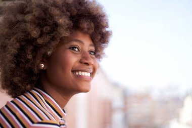 Afro saçlı mutlu siyah kadın gülümsüyor. Neşeli Afrikalı Amerikalı kız açık havada izliyor. Pencereden dışarı bakan kadın portresi. Boşluğu kopyala.