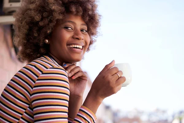アフロの髪をした幸せな若い黒人女性がカメラの笑顔を見てポーズ 陽気なアフリカ系アメリカ人の少女が屋外でコーヒーを飲んでいます 肯定的な表現を持つ女性の肖像画 スペースのコピー — ストック写真