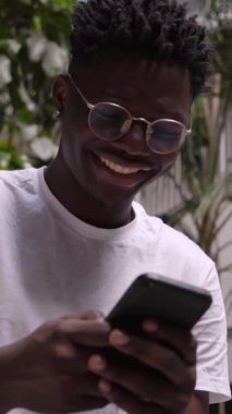 İşten çıkarken cep telefonu kullanan mutlu bir zencinin dikey videosu. Gülümseyen Afrikalı Amerikalı çocuk sosyal medyaya mesaj atıyor ve kontrol ediyor. Çevrimiçi bir uygulamaya bağlanan neşeli bin yıllık.