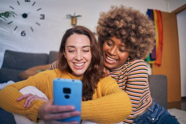 Evdeki oturma odasında gülümseyen ve cep telefonu kullanan mutlu eşcinsel kadın eşcinsel evlilik portresi. Birlikte hayatın tadını çıkaran çok ırklı lezbiyen partner ilişkisi. LGBT çalışanları. 