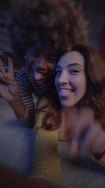 Dikey genç çok ırklı LGBT partneri içeride kameraya komik el sallıyor. Mutlu bir şekilde gülen iki lezbiyen kadının selfie 'si. Homoseksüel çift ilişkisi. İnsanlar geceleri aşktan zevk alıyorlar.