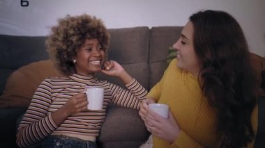 İki mutlu arkadaş oturma odasında oturmuş sohbet ediyor gülümsüyor ve bardakta sıcak bir şeyler içiyor. Hafta sonu rahatlama ve rahatlama aktiviteleri kavramı. Kadın çiftlerde rahatlama ve boş zaman. 