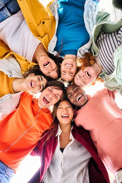 Vertical large multiracial group of smiling young people standing hugging looking at camera. Group of cheerful people in circle looking down. Low angle view. Copy space.