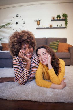Dikey iki mutlu arkadaş oturma odasında halıya uzanmış kameraya bakarak gülümsüyor. Hafta sonu rahatlama ve rahatlama aktiviteleri kavramı. Dinlen ve kız arkadaşlarınla vakit geçir.. 